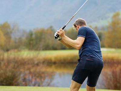 Man in blue outfit swinging a golf club on a lush golf course with mountains in the background on a clear day.