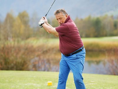 A man in a maroon shirt and blue pants swings a golf club on a grassy field with trees and a lake in the background.