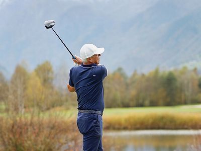 Golfer in blauer Kleidung und weißer Kappe schwingt auf einem Golfplatz, im Hintergrund Berge und Wald.