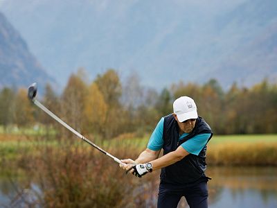 Person swinging a golf club on a course with mountains and autumn foliage in the background.
