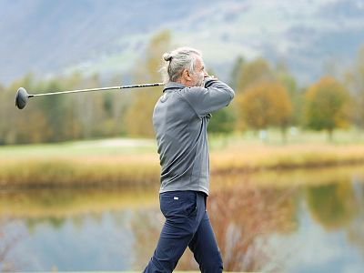 Older man with bun golfing on scenic course with autumn trees and mountains in the background, wearing gray jacket and pants.