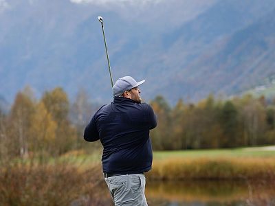 A golfer in motion swings at a course surrounded by trees and mountains, illustrating a serene outdoor sports setting with autumn colors.