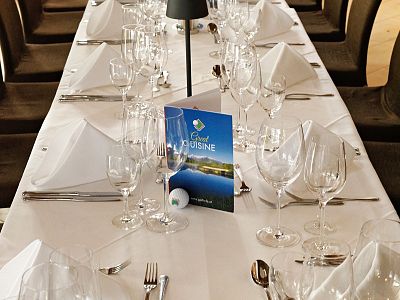 A formal dining table set with white tablecloth, folded napkins, wine glasses, and cutlery. A menu card stands upright in the center of the table.