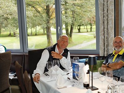 Mehrere ältere Männer sitzen an einem festlich gedeckten Tisch in einem Restaurant, mit großen Fenstern im Hintergrund, durch die ein grüner Garten zu sehen ist.
