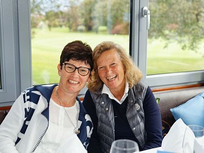 Two women smiling at a table inside, with a garden visible through the window. They are wearing casual attire and appear to be enjoying their time.