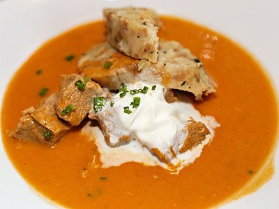 A bowl of goulash soup with meat chunks, creamy sauce, and sliced dumplings garnished with herbs, served in a white dish.