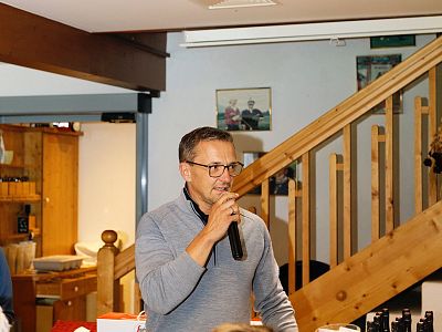 A man in glasses and a sweater speaks into a microphone indoors. Behind him is a wooden staircase, and the setting looks like a cozy room.