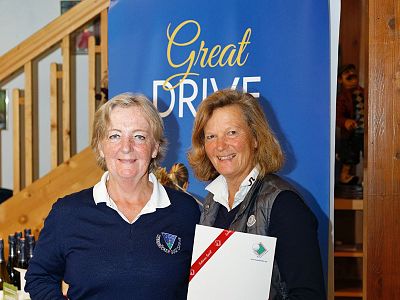 Two women smiling with a certificate in front of a "Great Drive" banner, likely at a golf event.