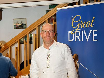 Man in white shirt at a 'Great Drive' event, standing next to a blue banner. Wooden staircase and frame pictures in the background.