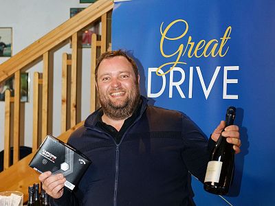 Man smiles holding a wine bottle and gift box in front of blue backdrop reading "Great Drive." Wooden staircase in background. Casual event atmosphere.