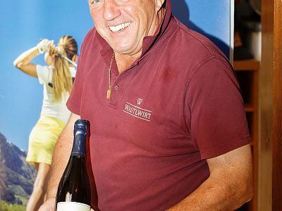 Man in maroon shirt holds a bottle, smiling. Background shows a golfing poster with a woman in action. Casual indoor setting.