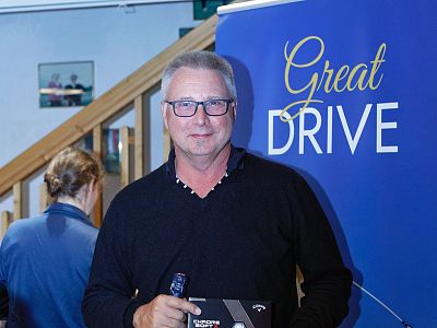 A man with glasses in front of a blue sign reading 'Great Drive', indoors, holding an award. Another person is seen from behind in the background.