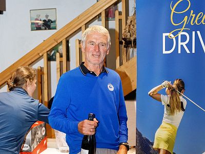 Man in blue sweater holding bottle at event with golf banner in background.