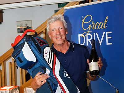 An older man smiling, holding a golf bag and a wine bottle. Behind him is a sign with "Great Drive" written on it, in an indoor setting.
