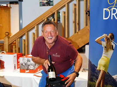 A smiling man holds a champagne bottle, surrounded by event gifts and a golf-themed poster at an indoor gathering.