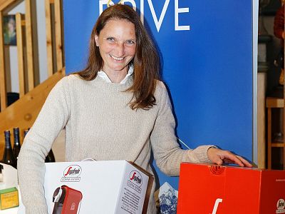Frau in beigem Pullover hält zwei bunte Boxen und lächelt. Im Hintergrund ist ein blaues Poster zu sehen. Verschiedene Flaschen stehen auf einem Tisch.