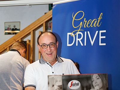 A man smiling, standing in front of a 'Great Drive' sign, holding an item. A staircase and another person are partially visible in the background.