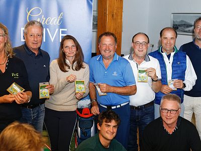 A group of nine smiling people, some holding booklets, pose indoors in casual attire. A banner reads "Great Drive." They appear to be at an event.
