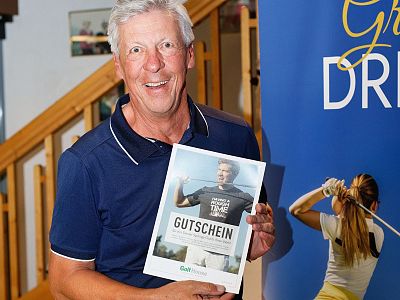 Ein lächelnder Mann hält einen Golf-Gutschein in der Hand. Er steht vor einem Roll-up-Banner, das Golf-Themen zeigt, in einem Innenraum.