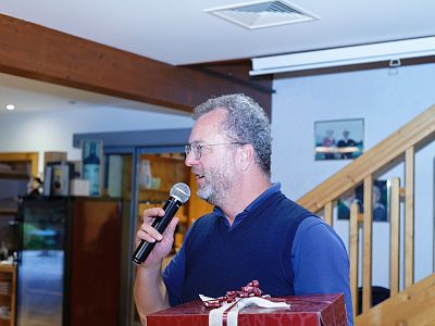 A man with glasses holds a microphone and a gift, speaking in a wood-decorated room, suggesting a social event.