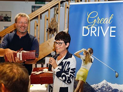 A man and woman holding gifts at a golf event. A banner with "Great Drive" and a golfer in action is in the background.