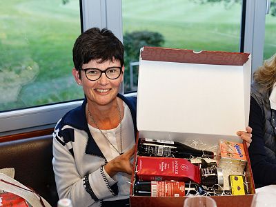 A woman with short dark hair and glasses holds an open gift box with assorted items like cosmetics and snacks. She sits by a window showing green scenery.