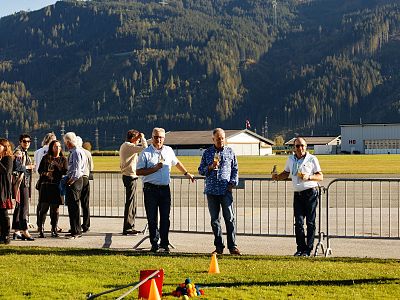 People at an outdoor event stand near a railing with scenic mountain views and clear blue skies, creating a backdrop for a relaxed gathering under the sun.