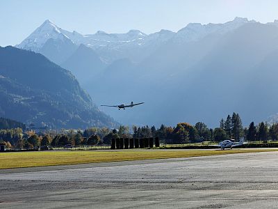 A small plane flying over green fields with tall, snow-capped mountains and a clear sky in the background.