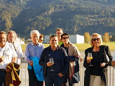 A group of people at an outdoor event with drinks, against a mountain backdrop, enjoying sunny weather.
