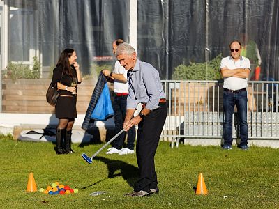 A man is playing mini golf on a grassy field with scattered cones and colorful balls. Onlookers stand in the background, suggesting a casual outdoor event.