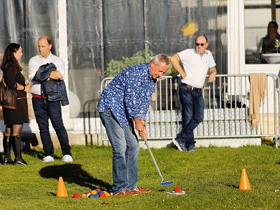 Man in floral shirt plays mini-golf on grass, others watch nearby. Cones and balls are on the ground.