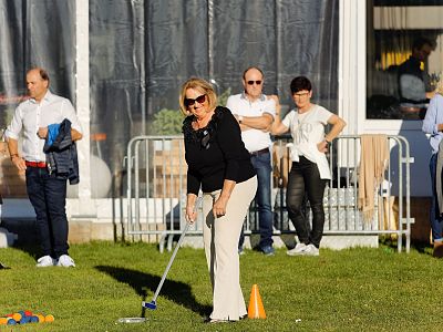 A woman playing mini-golf outdoors, surrounded by onlookers; sunny weather with a grassy background and nearby spectators in casual attire.
