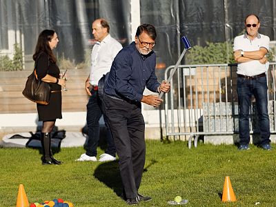 A man swings a golf club on a grass field during a gathering, with a few people observing in the background, including a woman and two men.