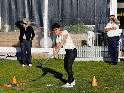 A woman swings a golf club on grass near cones and colorful balls, with three people watching in the background. Sunny outdoor setting.