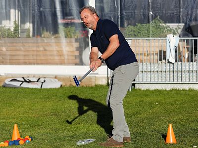 Person playing croquet on grass, focused on hitting ball with stick. Colorful balls and orange cones scattered in background.