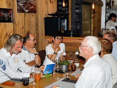 Several individuals dining in a rustic restaurant, sitting around a table with drinks and menus, amidst wooden furnishings and warm lighting.
