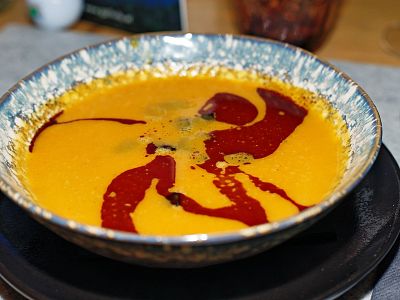 A bowl of bright orange soup with red swirls on top, placed on a blue and white patterned plate on a gray placemat.