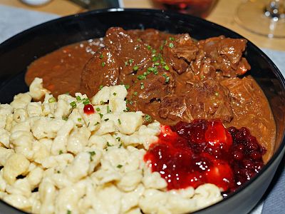 A plate with beef goulash, spaetzle noodles, and a side of lingonberry sauce garnished with herbs, presented on a dining table.