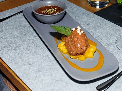 A dessert plate with chocolate mousse, a garnish leaf, mango slices, and a bowl of sauce, elegantly presented on a long, gray plate.