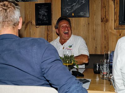 A group of people sitting at a table in a cozy room with wooden walls. They are engaged in conversation, and one person, wearing a white shirt, is laughing heartily.