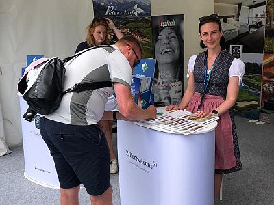 Man in casual attire signs form at a tourism booth with brochures, while woman in traditional dress stands nearby, smiling at an exhibition.