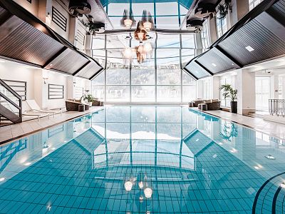 Spacious indoor pool with large windows and a skylight, offering natural light. Surrounding lounge chairs and plants create a relaxing atmosphere.