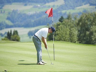Man putting on a golf course in sunny weather, surrounded by greenery and distant hills, aiming for the hole near a red flag.