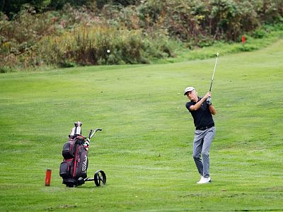 A golfer in mid-swing on a lush green course, with a golf bag nearby. The scene is bordered by trees and shrubs, providing a natural setting.