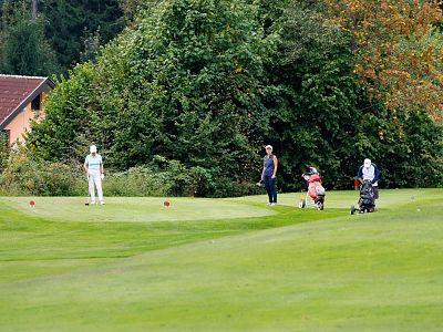 Golfers playing on a lush green course, surrounded by trees, with a house partially visible. A golfer near bags and others are on the course.