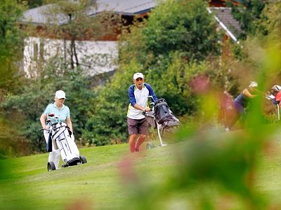 Two golfers push their equipment trolleys on a green course, surrounded by trees and a small building in the background.