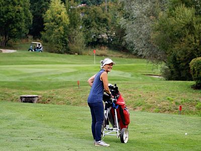 Woman in blue sportswear smiling on a golf course, holding a golf trolley with clubs. Lush green fairway and trees in the background. Another golfer is visible.