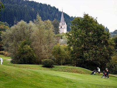 Golfers on a course surrounded by trees, with a church tower and forested hills in the background.