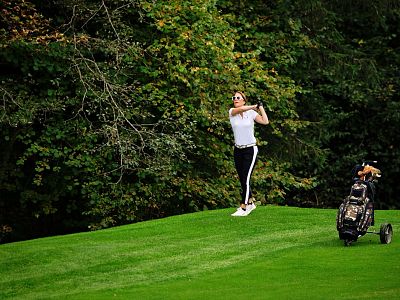 Person golfing on a green course surrounded by trees, with a golf bag on wheels nearby.