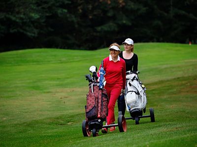 Two women on a golf course, each with a golf cart, walking on the green. They are dressed in sporty attire and appear to be enjoying the day outdoors.